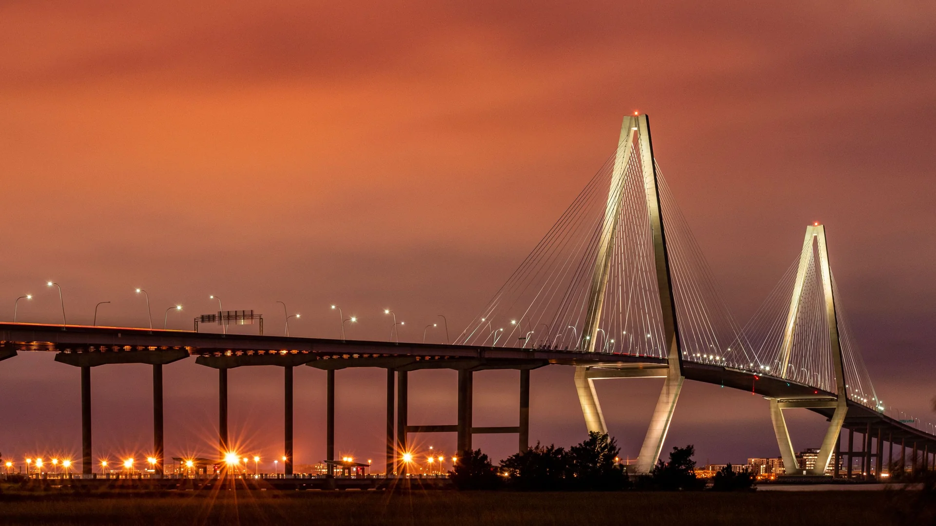 The Arthur Ravenel Jr. Bridge lit at dusk over Charleston Harbor, its two diamond towers glowing against a copper-orange sky